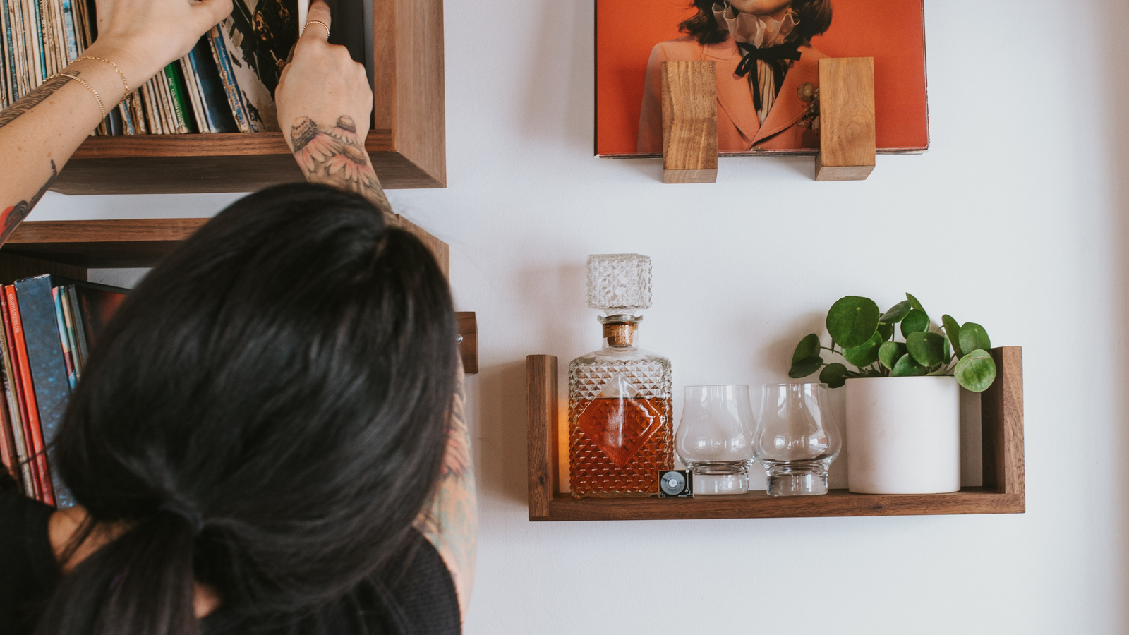 A woman looking through records in a walnut Wall Cube record shelf next to a floating shelf in walnut holding a decanter, some glasses, and a plant underneath a front facing record display shelf in walnut