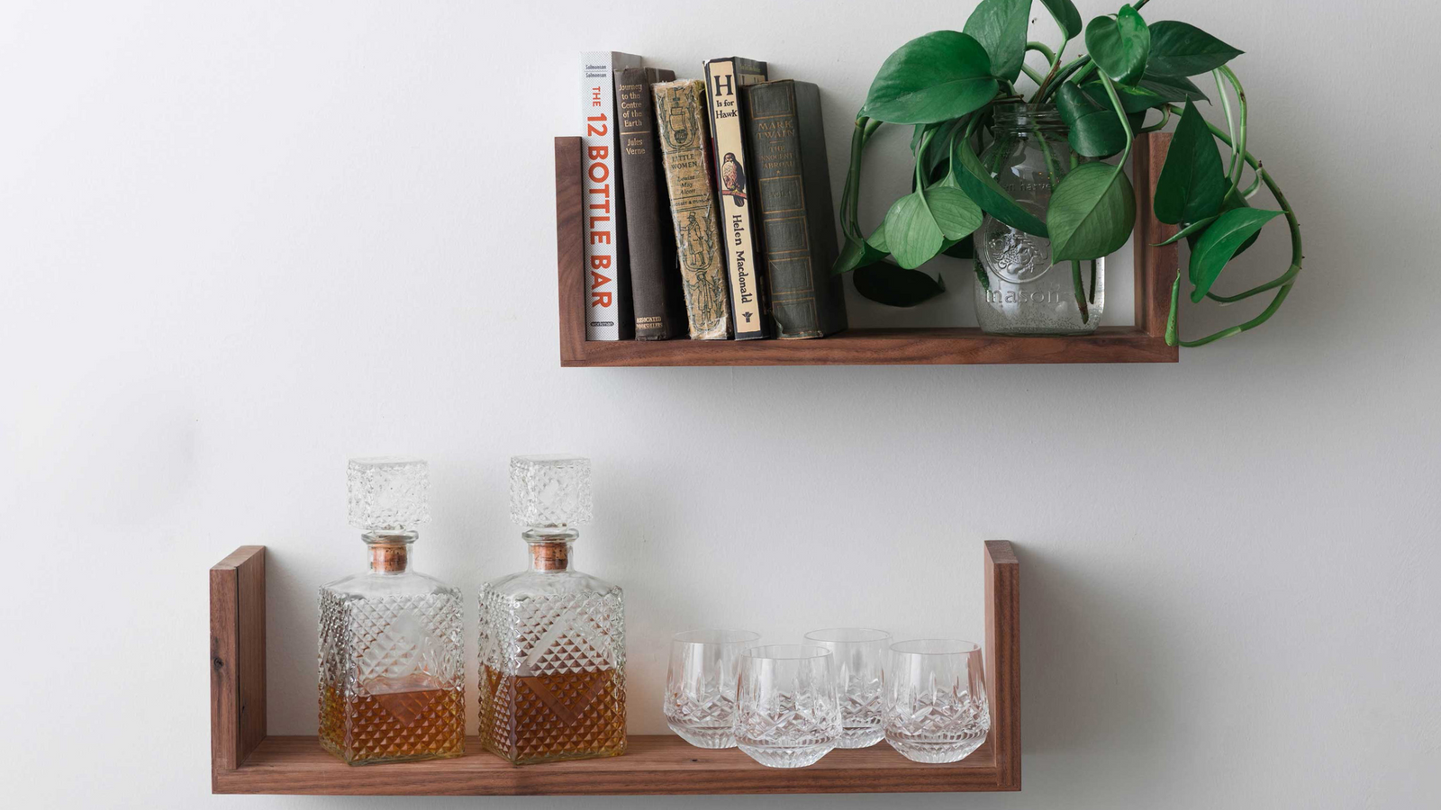 Floating book shelves with sidewalls mounted one above the other in varying sizes holding a glass decanter and 4 glasses as well as books and a plant